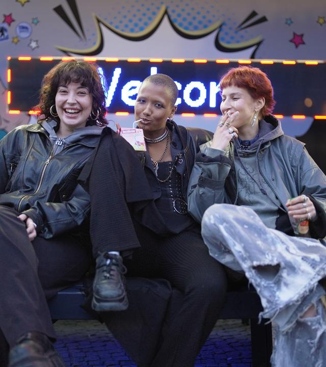 Drei junge Frauen vor einer Leuchtschrift, auf der „Welcome“ steht.