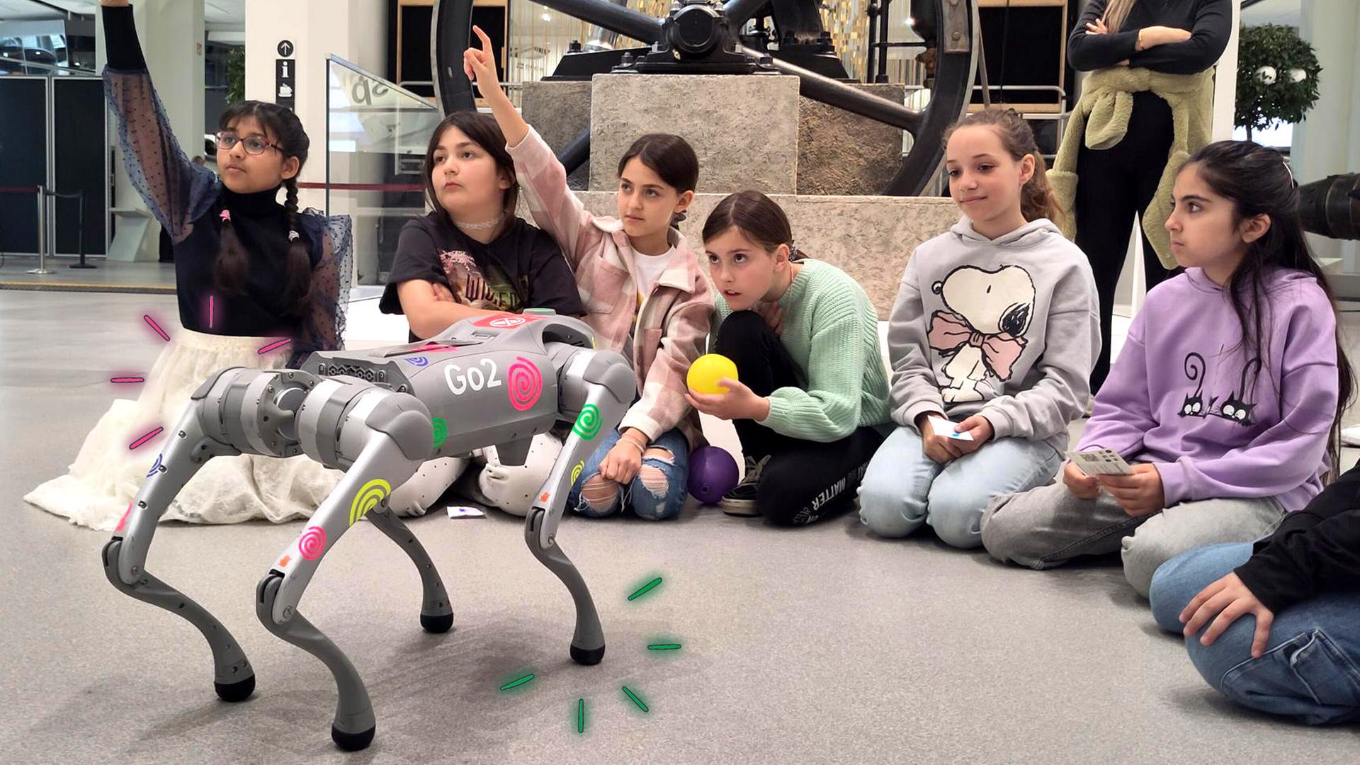 Kinder sitzen auf dem Boden, vor ihnen steht ein Roboterhund, zwei Mädchen melden sich