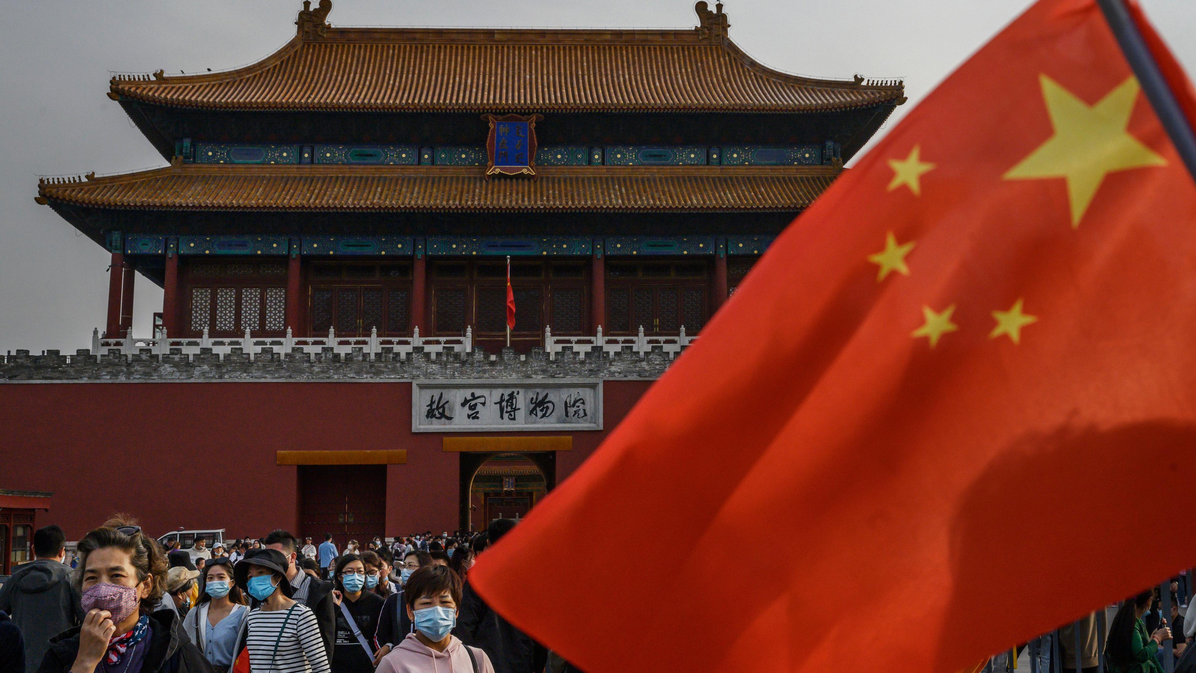 Eine chinesische Flagge, dahinter Menschen mit Mundschutz vor einem Gebäude der Verbotenen Stadt.