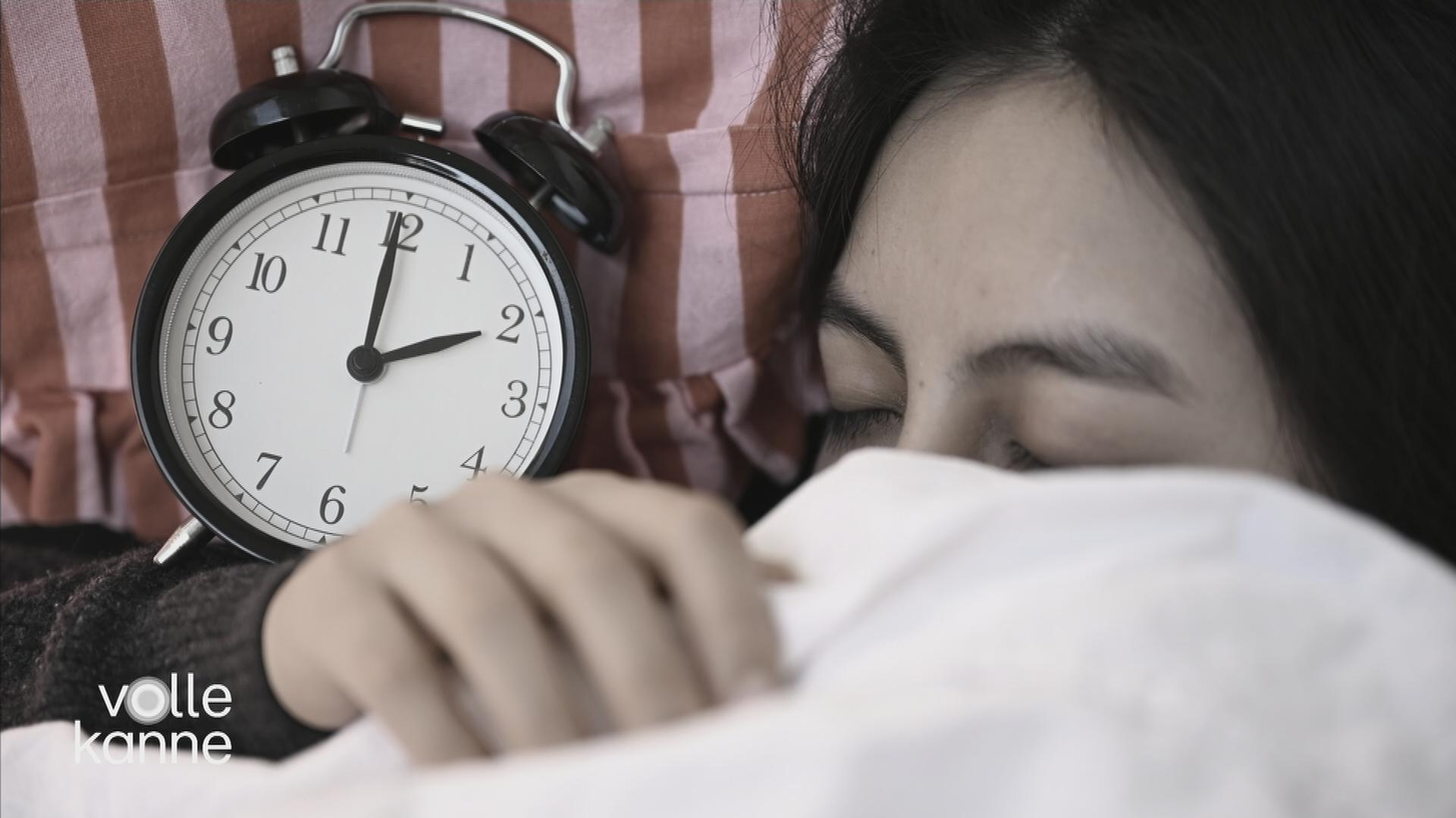 Eine Frau zieht sich die Decke über den Kopf. Neben ihr ist eine Uhr.