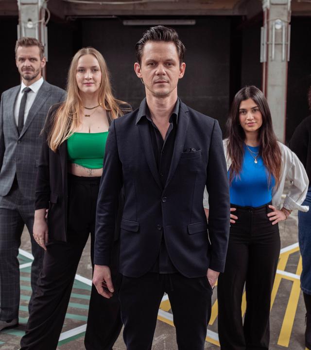 Sieben Menschen stehen auf einem Spielfeld und blicken in die Kamera.