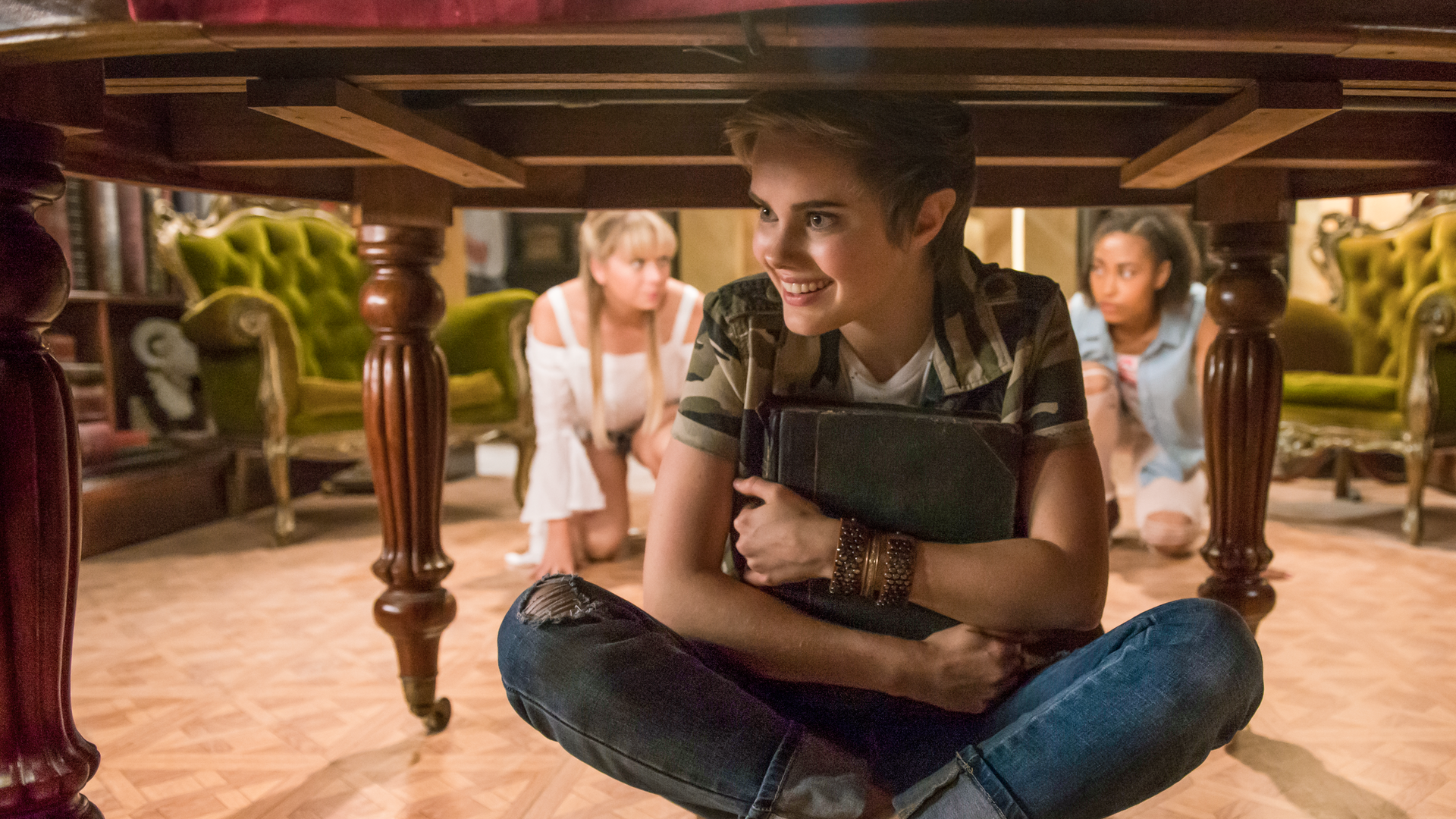 Imogen sitzt grinsend unter einem Tisch. Lily und Ruksy knien im Hintergrund.