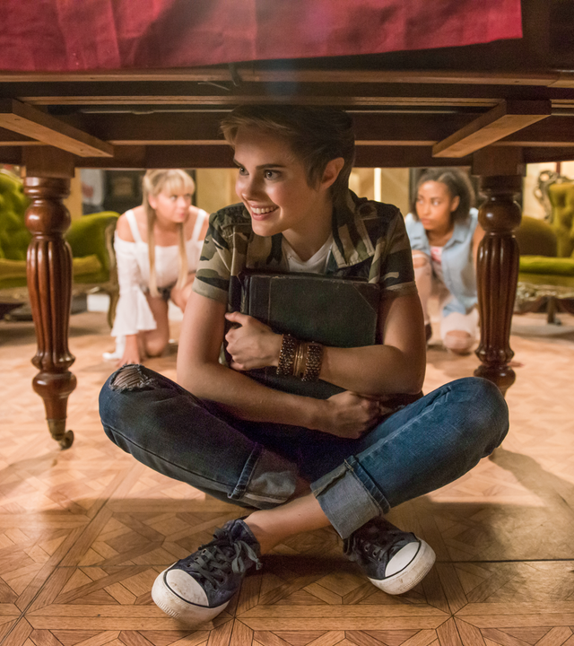 Imogen sitzt grinsend unter einem Tisch. Lily und Ruksy knien im Hintergrund.