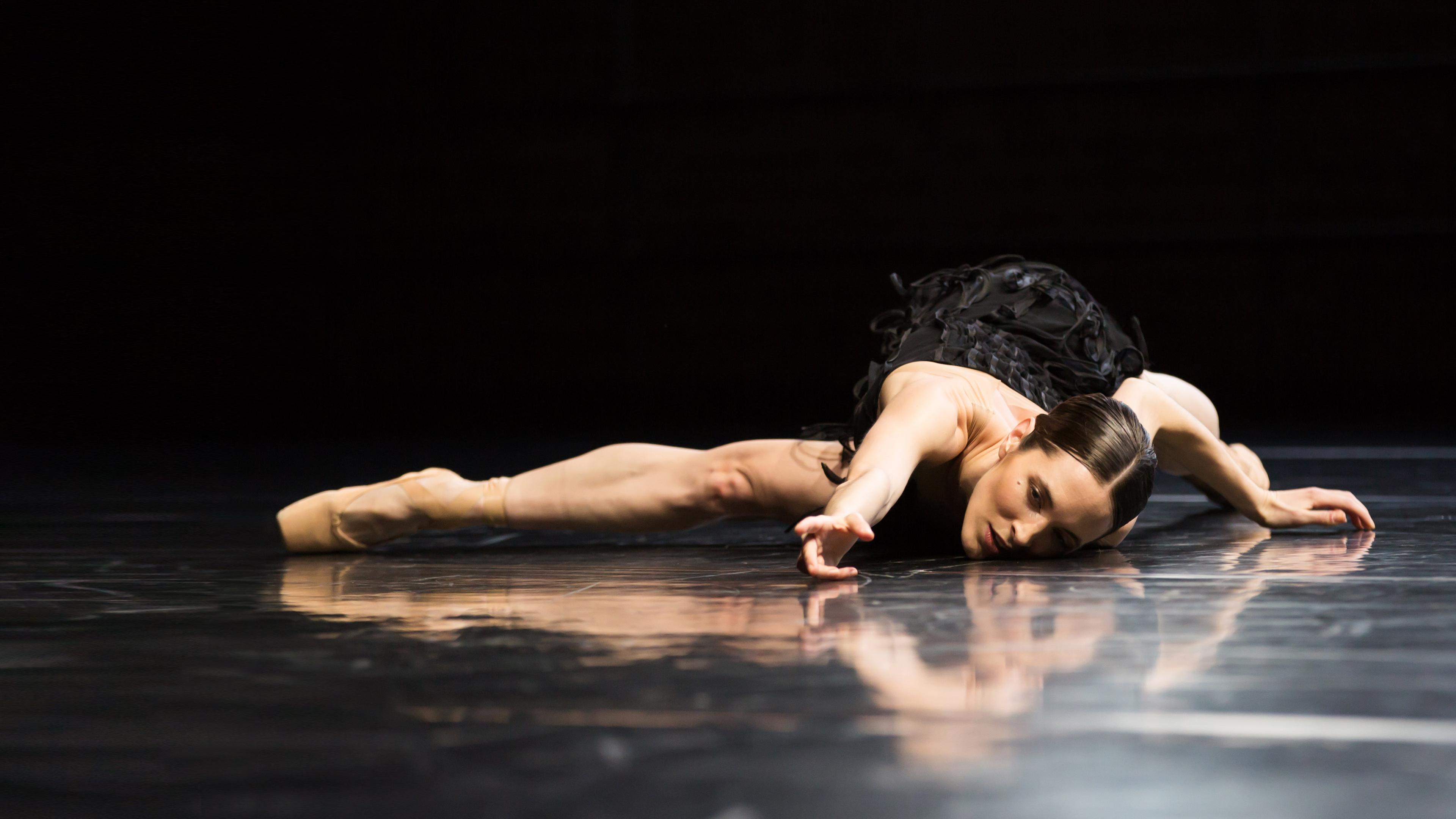 Die Tänzerin steht auf Zehenspitzen. Sie blickt nach oben und streckt die Arme in die Höhe. Sie trägt ein schwarzes Trikot mit Fransen.