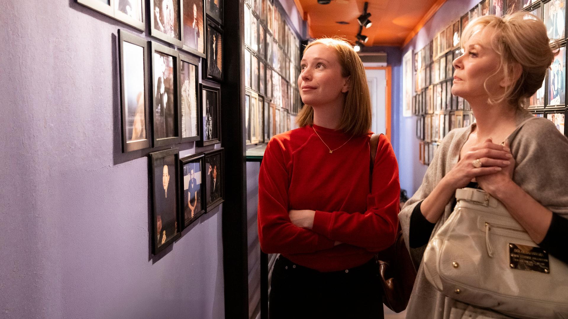 "Hacks - 1,69 Millionen": Ava ((Hannah Einbinder) und Deborah (Jean Smart) stehen in einem schmalen Flur, dessen Wände dicht mit gerahmten Fotografien bedeckt sind. Während Ava die Fotowand leicht lächelnd betrachtet, wirft Deborah einen nachdenklichen Blick darauf.