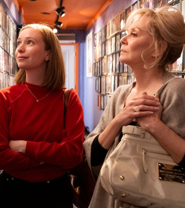 "Hacks - 1,69 Millionen": Ava ((Hannah Einbinder) und Deborah (Jean Smart) stehen in einem schmalen Flur, dessen Wände dicht mit gerahmten Fotografien bedeckt sind. Während Ava die Fotowand leicht lächelnd betrachtet, wirft Deborah einen nachdenklichen Blick darauf.
