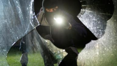 Einbrecher mit Maske und Taschenlampe leutet durch zebrochene Glasscheibe