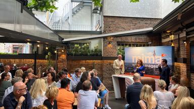  Jahrespräsentation der ZDF Werbefernsehen GmbH 2024 in Frankfurt: Hans Joachim Strauch, Mirjam Meinhardt und Andreas Wunn stehen an einem Tisch mit Mikrofonen in der Hand. Im Vorderegrund das Publikum, das an Biertischen Platz genommen hat.