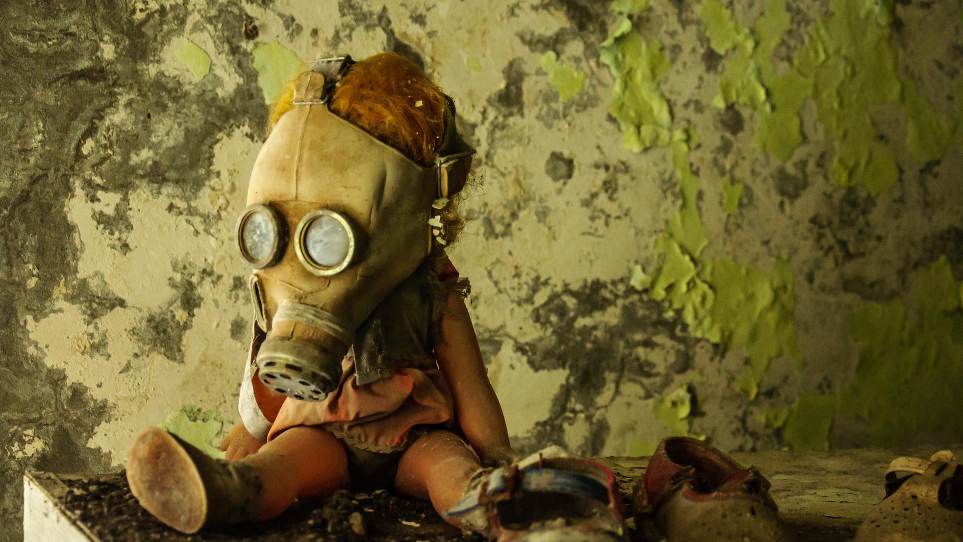 "Tschernobyl - Die Katastrophe": Eine Kinderpuppe trägt eine Gasmaske. Sie sitzt vor einer abbröckelnden Tapete auf einer schmutzigen Fläche. Daneben steht ein Paar Kinderschuhe.
