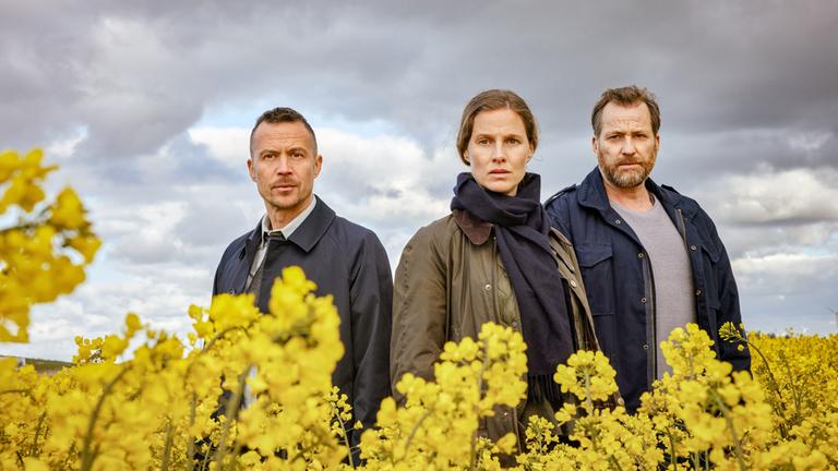 "Ostfriesenfluch": Rupert (Barnaby Metschurat,), Ann Kathrin Klaasen (Picco von Groote) und Frank Weller (Christian Erdmann) stehen mitten in einem gelben Rapsfeld etwas versetzt nebeneinander. Sie sind nur ab dem Oberkörper aufwärts zu sehen.