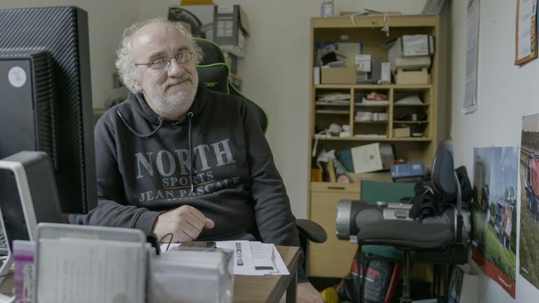 "Zwischen Not und Neustart": Harald (59) sitzt in seinem kleinen Redaktionsbüro vor dem Computer. Im Hintergrund ist ein Regal voller kleinerer Gegenstände und Unterlagen zu sehen.