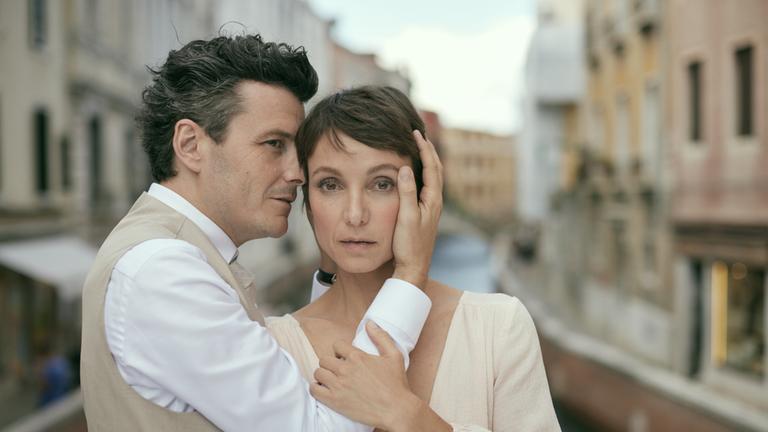 "Ewig Dein": Über einem Kanal in Venedig stehen Hannes (Manuel Rubey)  und Judith (Julia Koschitz) erhöht vor den alten Häuserfluchten, die sich rechts und links den Kanal entlangziehen. Hannes ist von der Seite zu sehen, Judith blickt direkt und starr in Richtung Kamera. Hannes hält ihren Kopf mit beiden Händen und zieht sie zu sich, während Judith eine Hand auf Hannes Arm gelegt hat.