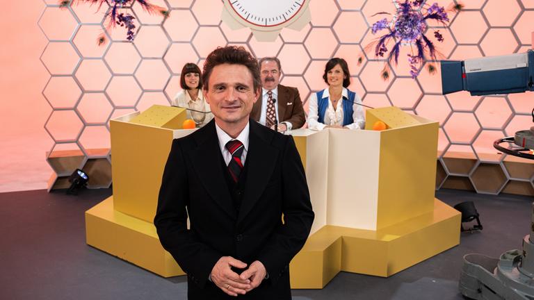 "Rosenthal": 70er Jahre: Hans Rosenthal steht im schwarzen Anzug vor einer Wabenwand im Studio. Hinter ihm sitzt seine Ratejury, Mady Riehl (Theresia Wald), "Ekki" Fritsch (Timo Dierkes) und Brigitte Xander (Katharina Völkl), an Pulten.