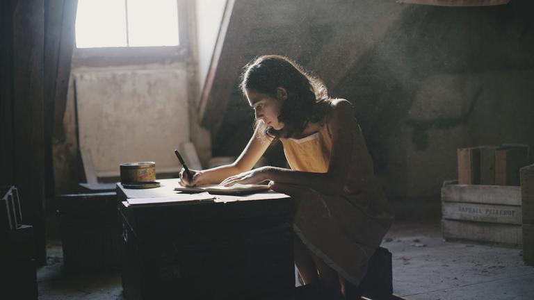 "Anne Frank. Tagebuch einer Jugendlichen": Das Bild zeigt ein Mädchen, das an einer Holzkiste sitzt und diese als Schreibtisch benutzt. Es hält einen Stift in der Hand und schreibt konzentriert in ein Buch. Auf der Kiste liegt eine alte Blechdose. Der Hintergrund ist unaufgeräumt und dreckig, durch ein Fenster fällt Licht ins Zimmer.