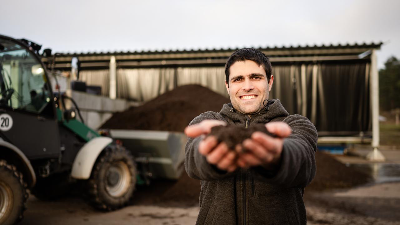"Kreative Köpfe, große Träume - Nachhaltig wirtschaften": Florian steht vor einer Halle neben einem Traktor. Er hat die Arme ausgestreckt und hält in beiden Händen seinen Dünger. Er schaut lächelnd in die Kamera.