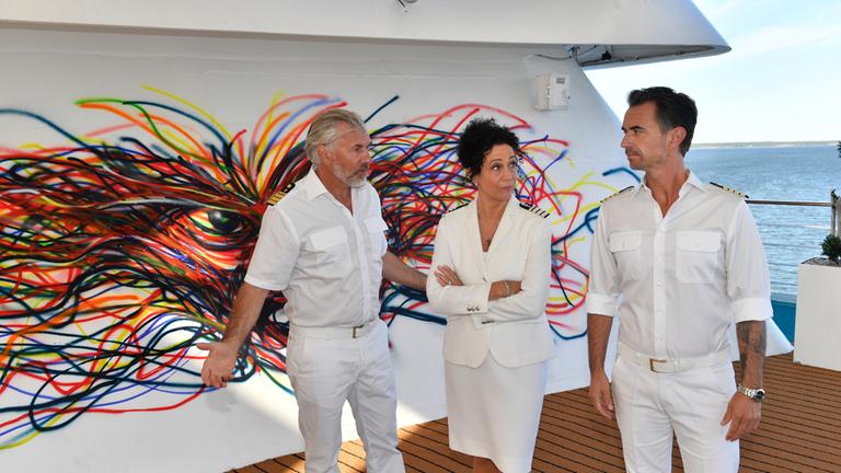 "Das Traumschiff: Miami": An Deck des Schiffs: Mit fragenden Blicken stehen Kapitän Max Parger (Florian Silbereisen), Hanna Liebhold (Barbara Wussow) und Staff-Kapitän Martin Grimm (Daniel Morgenroth) zusammen und unterhalten sich. Hinter ihnen an der Wand prangt ein buntes Graffiti.