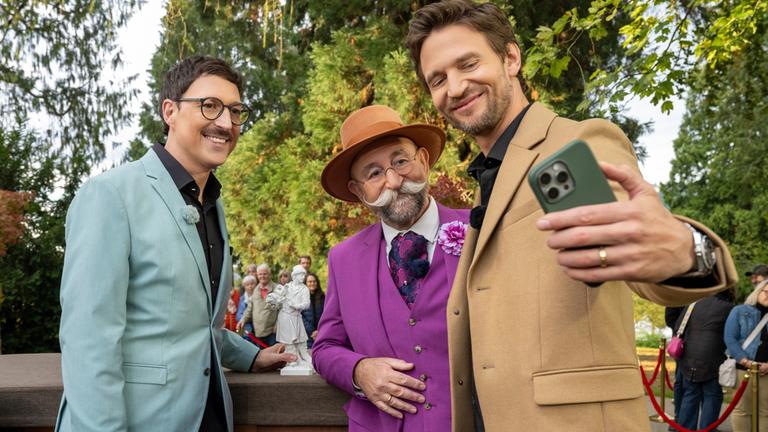 "Bares für Rares - Deutschlands größte Trödelshow": Colmar Schulte-Goltz, Horst Lichter und August Wittgenstein stehen dicht nebeneinander am Expertisenpult und lachen in Wittgensteins Handy, das dieser zum Selfie ausgestreckt in der linken Hand hält. Im Hintergrund steht das Publikum vor Bäumen.