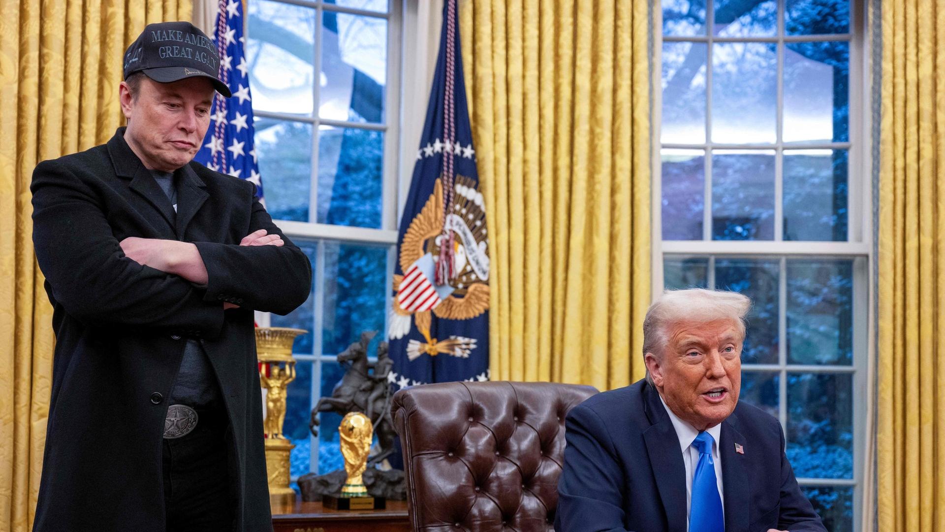 "Putins Helfer - Trump und Musk in Diensten des Kreml?": Elon Musk hört stehend und mit verschränkten Armen zu, während Donald Trump, im braunen Ledersessel im Oval Office sitzend, spricht. Im Hintergrund sind zwei Fenster mit goldfarbenen Vorhängen, dazwischen hängt die US-Fahne.