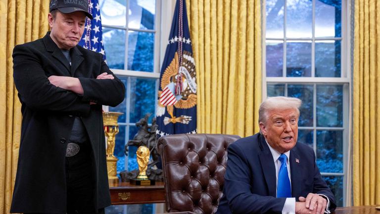"Putins Helfer - Trump und Musk in Diensten des Kreml?": Elon Musk hört stehend und mit verschränkten Armen zu, während Donald Trump, im braunen Ledersessel im Oval Office sitzend, spricht. Im Hintergrund sind zwei Fenster mit goldfarbenen Vorhängen, dazwischen hängt die US-Fahne.
