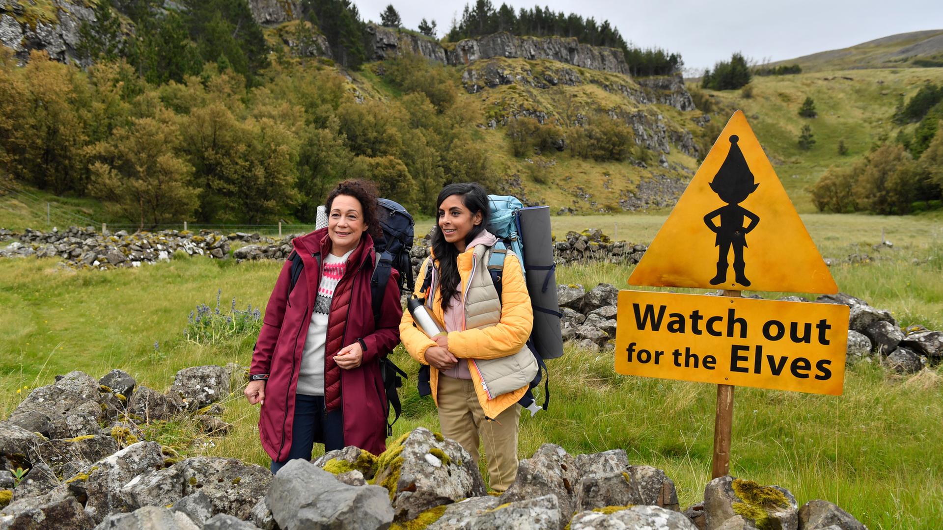 "Das Traumschiff: Island": In einer nordischen Naturkulisse: Auf einer grünen Wiese stehen Hanna Liebhold (Barbara Wussow) und Dr. Jessica Delgado (Collien Fernandes) nebeneinander und blicken lachend in die Ferne. Sie tragen dicke Jacken und Wanderrucksäcke. Dr. Delgado hält eine Thermoskanne in den Händen. Vor ihnen und auch im Hintergrund sind moosbewachsene Steine zu kleinen Mauern aufgetürmt. Neben ihnen steht ein gelbes Warnschild mit dem Abbild eines Elf und der Aufschrift "Watch out for the Elves".
