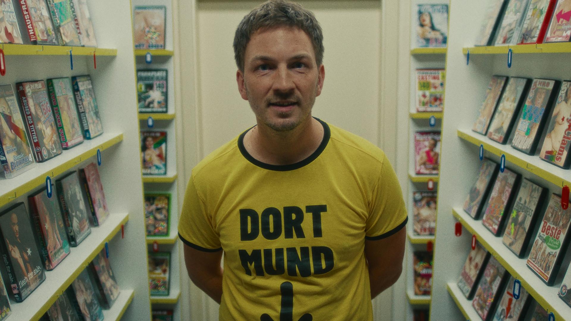 "Chabos - Warum liegt hier eigentlich Stroh?": Der erwachsene Peppi (Johannes Kienast) steht zwischen Regalen mit Porno-DVDs. Er trägt ein gelbes T-Shirt mit der schwarzen Aufschrift "Dort-Mund" und einem schwarzen Pfeil, der Richtung Schritt zeigt.