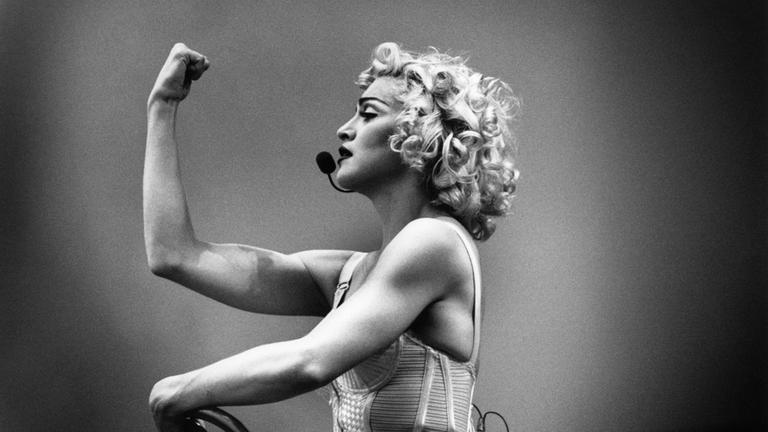"Becoming Madonna": Madonna trägt ein Headset und spannt in einer Power-Geste ihren Bizeps an.