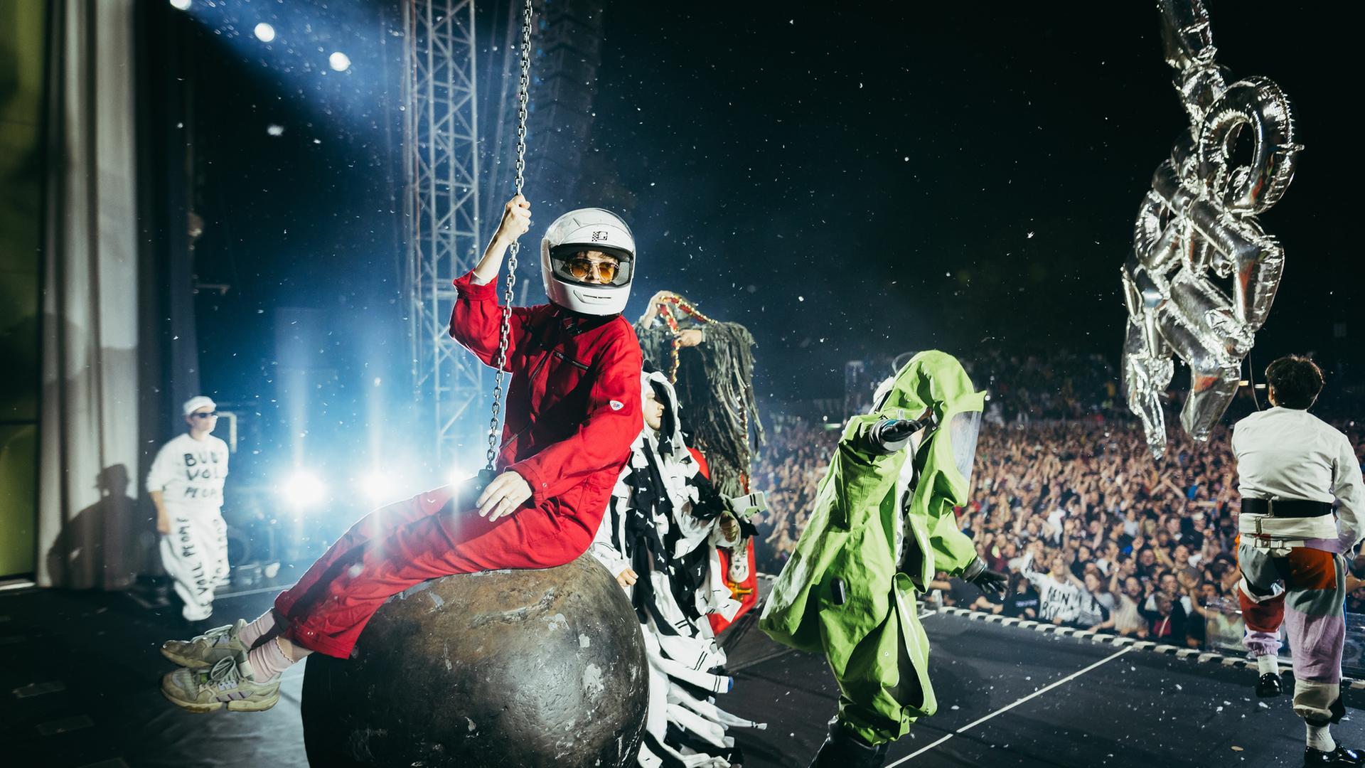 "Deichkind – Könnt ihr nochmal?": Das Bild zeigt eine dynamische Bühnenszene während eines. Im Mittelpunkt sitzt eine Person in einem roten Overall und mit weißem Helm auf einer großen Abrisskugel, die an einer Kette hängt. Weitere Personen in auffälligen Kostümen sind auf der Bühne verteilt: Eine trägt ein grünes Outfit mit langen Ärmeln und einer Art Maske oder Kopfbedeckung, eine andere ist in Weiß gekleidet mit schwarzem Text auf der Kleidung. Am rechten Bildrand ist eine weitere Person mit bunter Hose und weißem Oberteil teilweise sichtbar. Über der Bühne schwebt eine ballonartige Figur, möglicherweise aufblasbar oder metallisch glänzend. Im Hintergrund ist ein großes Publikum unter hellen Scheinwerfern und Bühnenlichtern zu sehen.