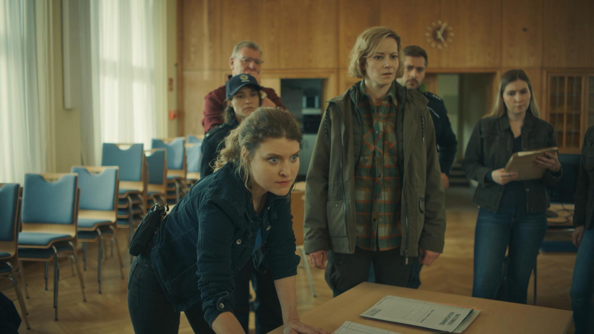 "Erzgebirgskrimi – Über die Grenze": Karina Szabo (Lara Mandoki) und Saskia Bergelt (Teresa Weißbach) stehen in einem Saal vor einem Tisch mit Ermittlungsergebnissen. Im Hintergrund stehen weitere Polizisten und Polizistinnen (Komparsen). Alle zusammen schauen konzentriert seitlich nach rechts.