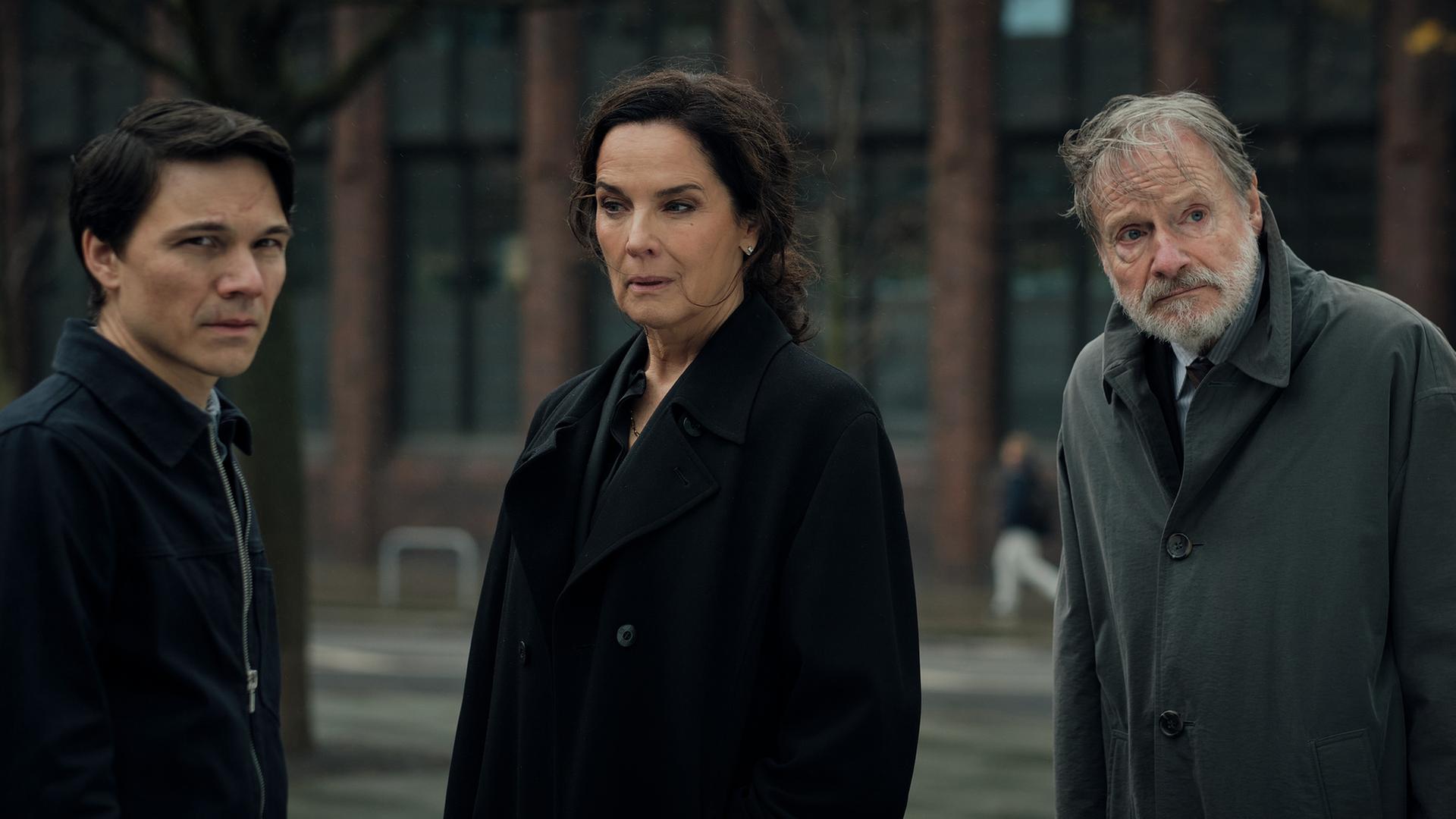 "Ein Fall für Conti – Der verlorene Sohn": Falk Klopfer (Sebastian Urzendowsky), Anna Conti (Désirée Nosbusch) und Friedrich Klopfer (Michael Wittenborn) stehen mit ernsten Gesichtsausdrücken nebeneinander. Hinter ihnen befinden sich eine Straße sowie ein Gebäude mit roter Backsteinfassade und hohen Fenstern.