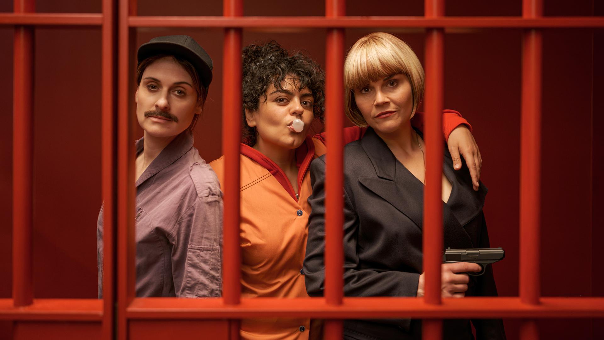 "Gar kein Geld macht auch nicht glücklich": Lesley (Julia Becker), Olivia (Sara Fazilat) und Kim (Katharina Wackernagel) stehen lässig nebeneinander in einem Raum mit knallroten Wänden. Vor ihnen sind rote Gitterstäbe, die die drei Frauen darin einsperren. Olivia formt mit dem Mund eine Kaugummiblase und legt ihren Arm um Kims Schultern. Diese hält eine Pistole in der Hand.