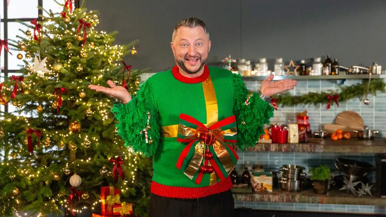 "besseresser: Die Tricks in Weihnachtsprodukten": Sebastian Lege trägt einen Weihnachtspulli mit großer Schleife und steht vor einem Weihnachtsbaum.