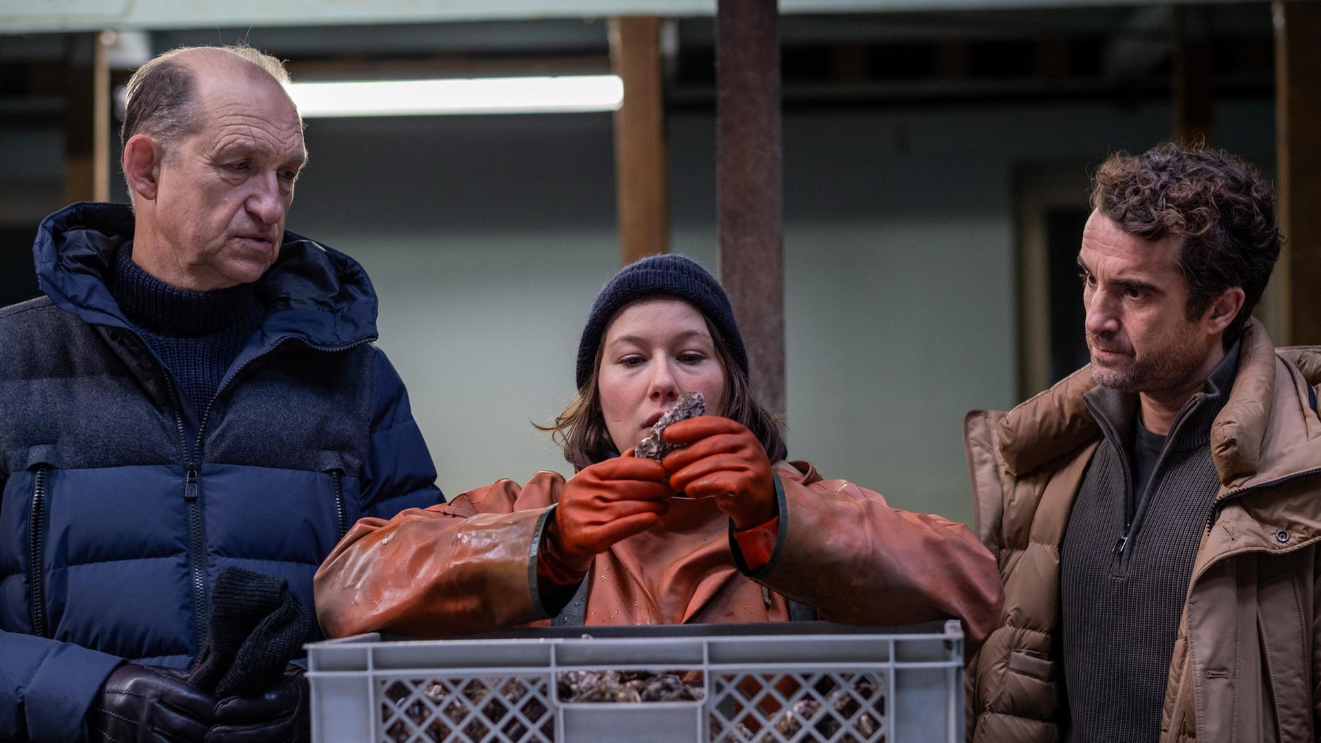  "Nord Nord Mord - Sievers und die stillen Austern": Tomke Bundis (Josefine Israel) beäugt eine Auster, die sie aus der vor ihr stehenden Kiste genommen hat. Carl Sievers (Peter Heinrich Brix) und Hinnerk Feldmann (Oliver Wnuk) stehen links und rechts neben ihr und schauen argwöhnisch.