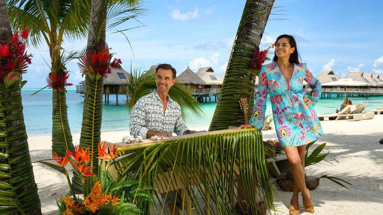"Das Traumschiff: Bora Bora": Am Sandstrand: Unter Palmen ist ein Keyboard aufgebaut, das mit einem Palmenwedel und Muscheln dekoriert ist. An diesem sitzt Kapitän Max Parger (Florian Silbereisen) und lächelt. Dr. Jessica Delgado (Collien Ulmen Fernandes) lehnt sich an das Keyboard und lächelt ebenfalls.
