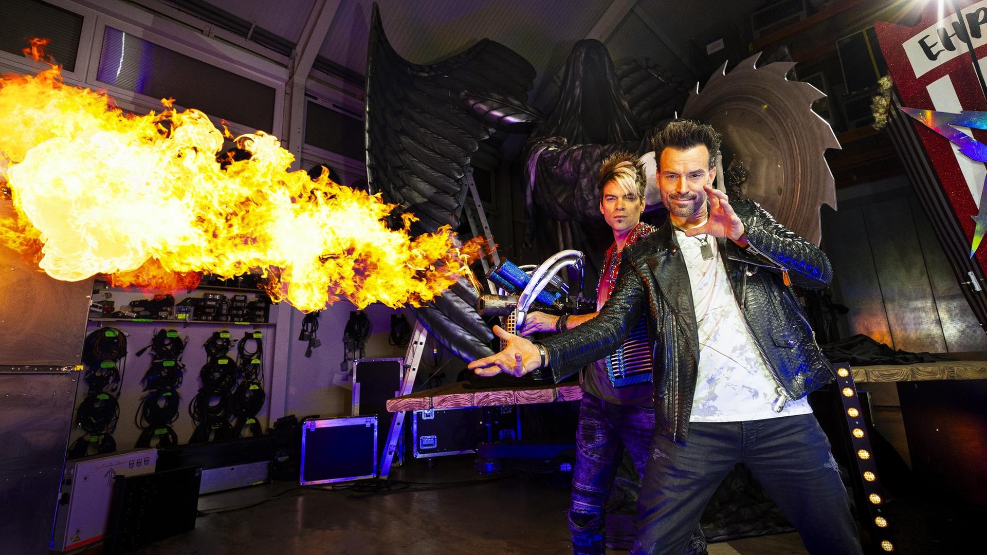 "Backstage - Ehrlich Brothers - Die Magie hinter der Diamonds-Show": Chris Ehrlich und Andreas Ehrlich stehen vor einer großen Säge. Chris Ehrlich hat einen Flammenwerfer in der Hand, dem seitlich eine große Flamme entweicht.
