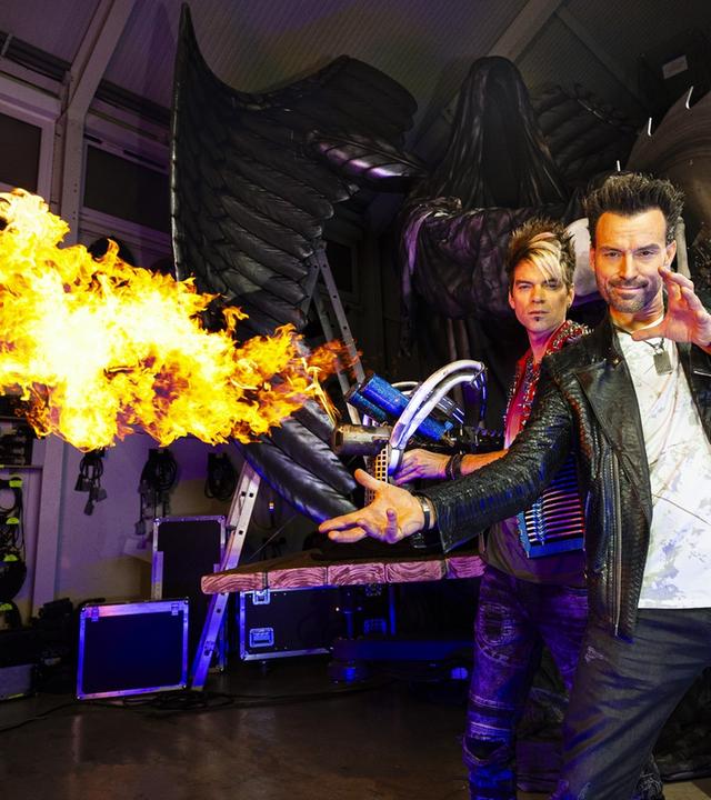 "Backstage - Ehrlich Brothers - Die Magie hinter der Diamonds-Show": Chris Ehrlich und Andreas Ehrlich stehen vor einer großen Säge. Chris Ehrlich hat einen Flammenwerfer in der Hand, dem seitlich eine große Flamme entweicht.