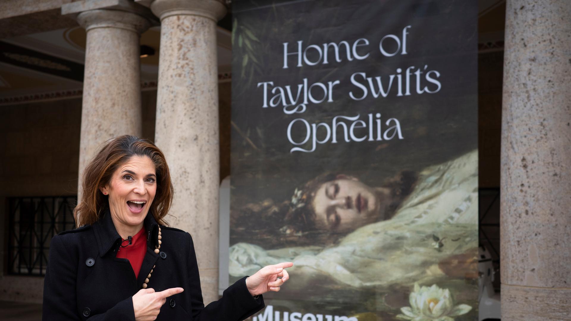 "WISO-Dokumentation: Kultur sucht Geld: Was wollen wir uns noch leisten?": Moderatorin Katty Salié steht am dem Eingang des Landesmuseums Wiesbaden vor einem Plakat mit dem Titel "Home of Taylor Swifts Ophelia". Sie schaut lächelnd in die Kamera und zeigt auf das Plakat.