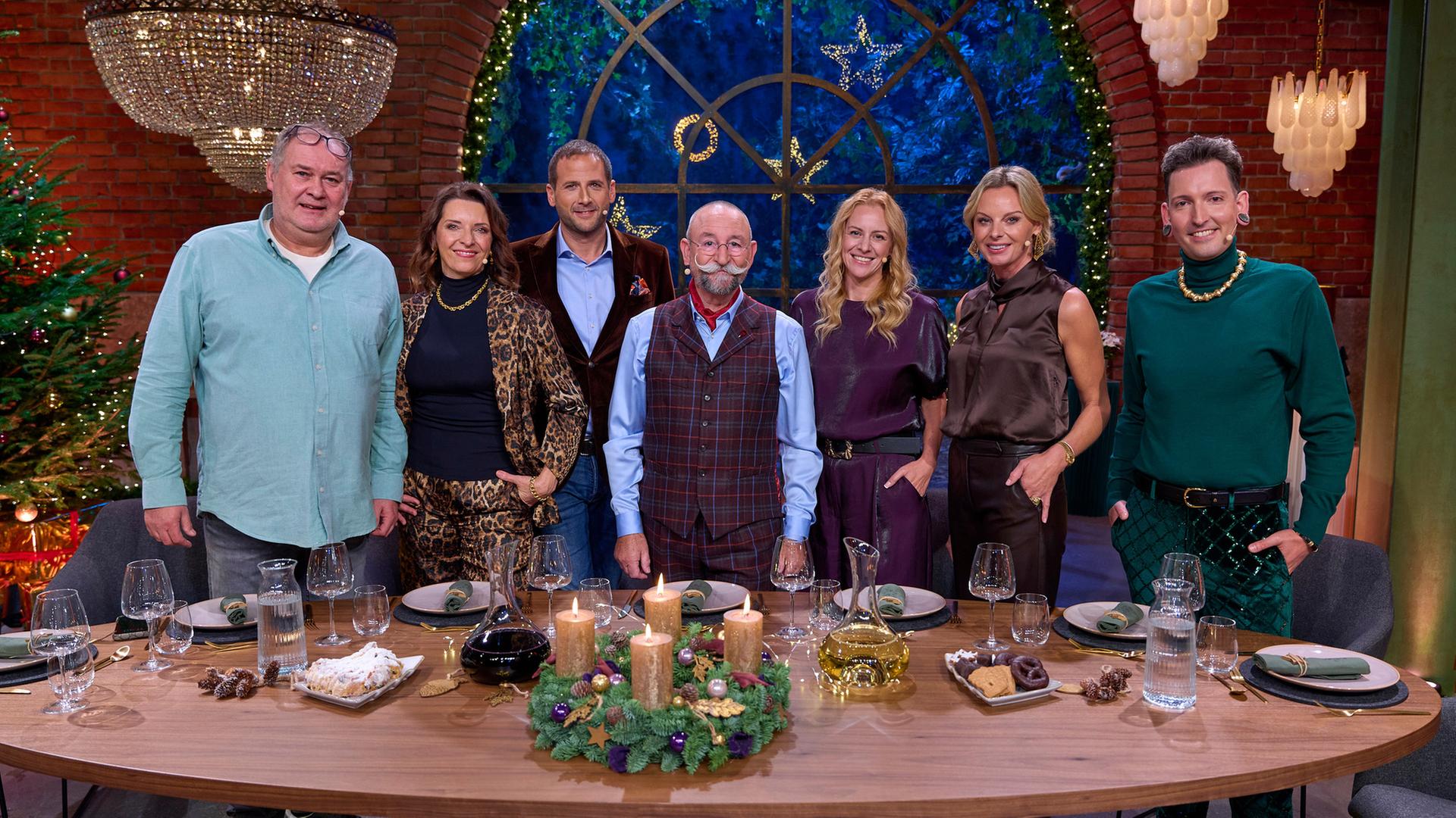 "Der "Bares für Rares"-Weihnachtsabend - mit Horst Lichter und Freunden": Walter Lehnertz, Wendela Horz, Julian Schmitz-Avila, Horst Lichter, Annika Raßbach, Susanne Steiger und Fabian Kahl stehen hinter einer festlich gedeckten Tafel und lächeln in die Kamera. Im Hintergrund ist ein großes geschmücktes Fenster sowie Weihnachtsdekoration zu sehen.