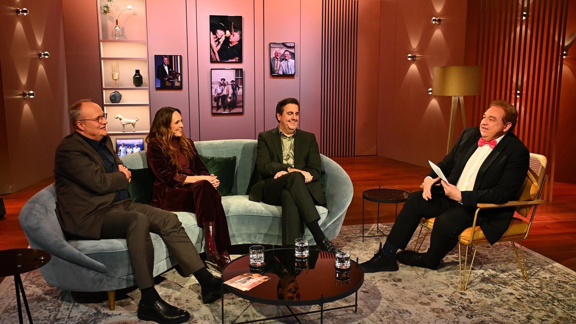 "Welke & Pastewka – Wiedersehen macht Freude!": Oliver Welke, Carolin Kebekus und Bastian Pastewka sitzen auf der Couch im Studio. Sie schauen alle drei zu Oliver Kalkofe, der rechts neben ihnen mit Moderationskarten in der Hand auf einem Stuhl sitzt.