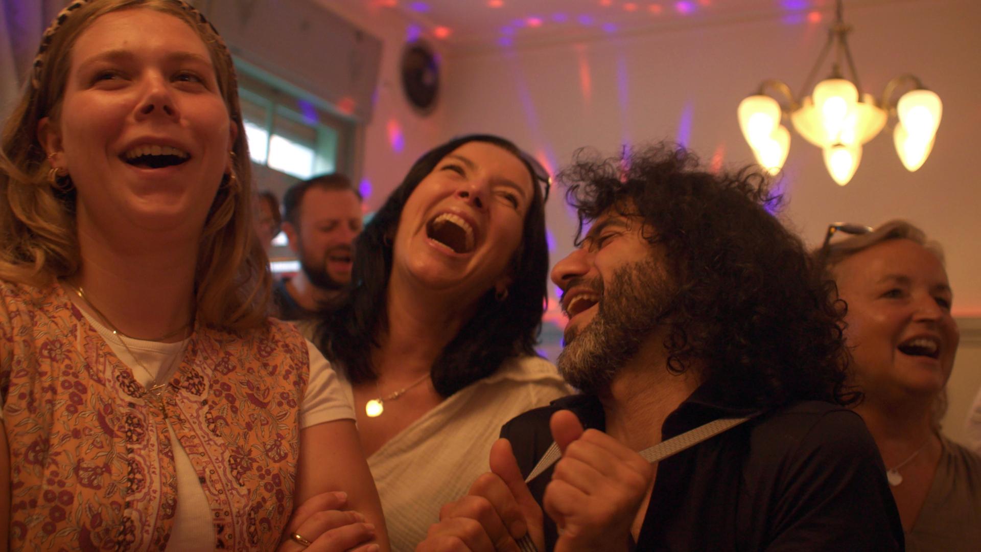 "Mehr als nur Musik - Die Kraft der Chöre": Zwei Frauen, ein Mann singen einander zugewandt in einem Raum. Im Bildhintergrund singen weitere Sänger und Sängerinnen.