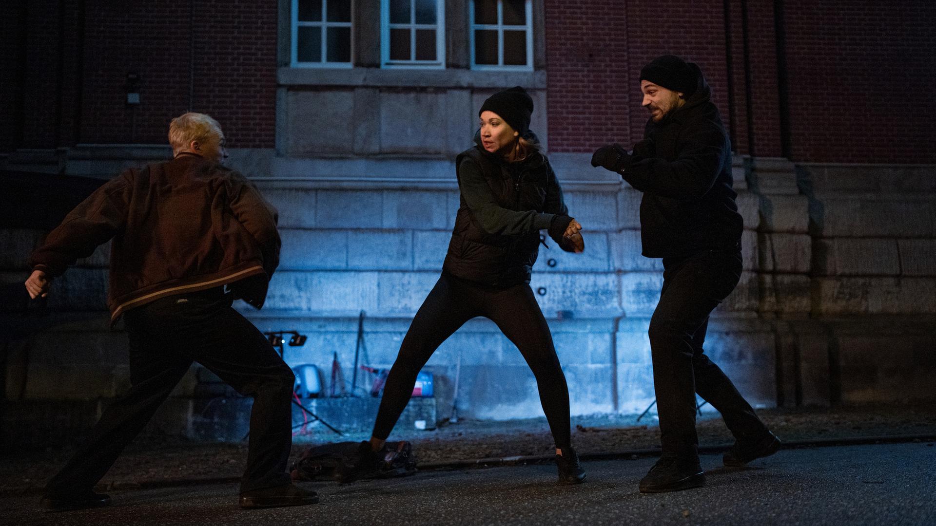 "Sarah Kohr – Großer Bruder": Nachts auf einer Straße vor einem größeren Haus, das indirekt beleuchtet wird, kämpft Sarah Kohr (Lisa Maria Potthoff) gegen Lars Rütger (Julius Nitschkoff) und Rainer Quentin (Boži Kocevski), die sie von beiden Seiten angreifen.