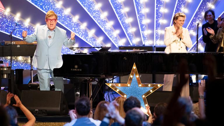 "An Evening with Elton John & Brandi Carlile": Elton John und die Sängerin Brandi Carlile stehen auf einer Bühne seitlich, bzw. hinter einem schwarzen Konzertflügel und blicken freudig ins Publikum.