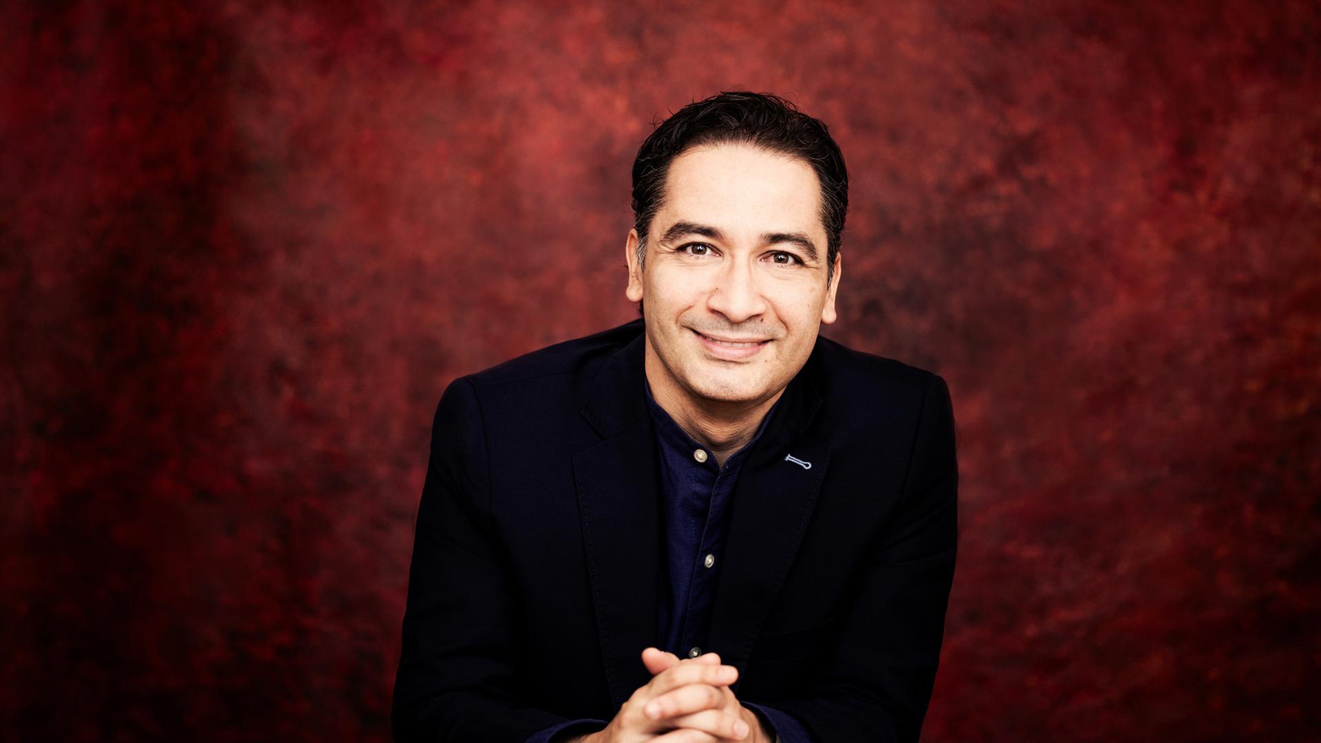 "Silvesterkonzert aus der Semperoper": Portraitaufnahme: Dirigent Andrés Orozco-Estrada steht vor einem rot-schwarz-marmorierten Hintergrund, seine Hände sind vor dem Körper gefaltet und er lächelt in die Kamera.