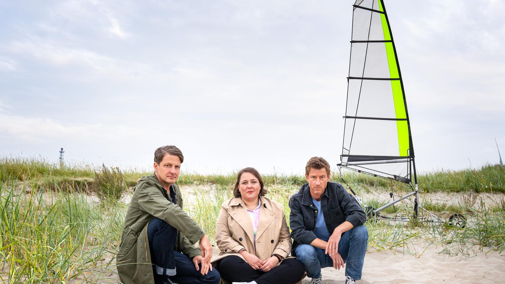 "Friesland - Geisterstunde": Wolfgang Habedank (Holger Stockhaus), Melanie Harms (Tina Pfurr) und Henk Cassens (Maxim Mehmet) sitzen im Sand einer Dünenlandschaft, im Hintergrund steht ein Strandsegler.