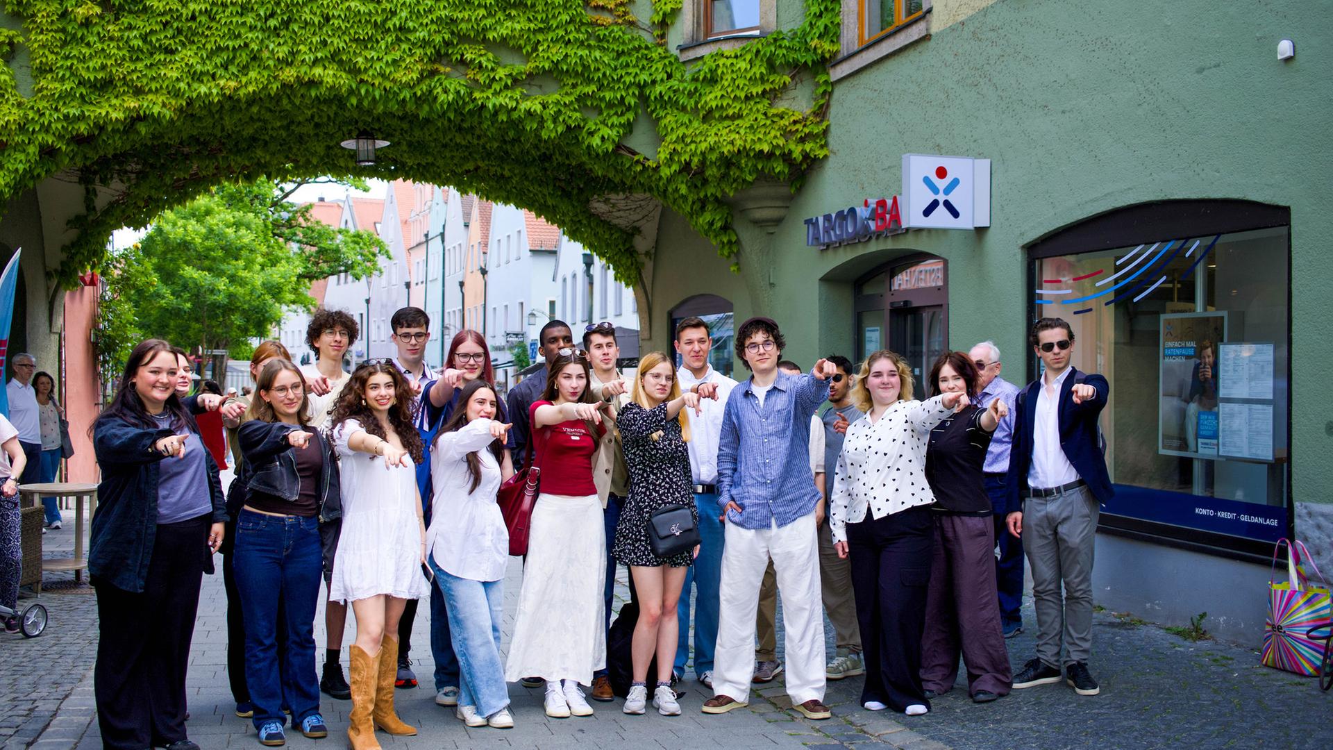 Bildbeschreibung: "plan b: Mut zur Versöhnung - Frieden statt Feindschaft": Eine Gruppe von jungen Menschen steht vor einem begrünten Torbogen. Sie alle zeigen nach vorne.