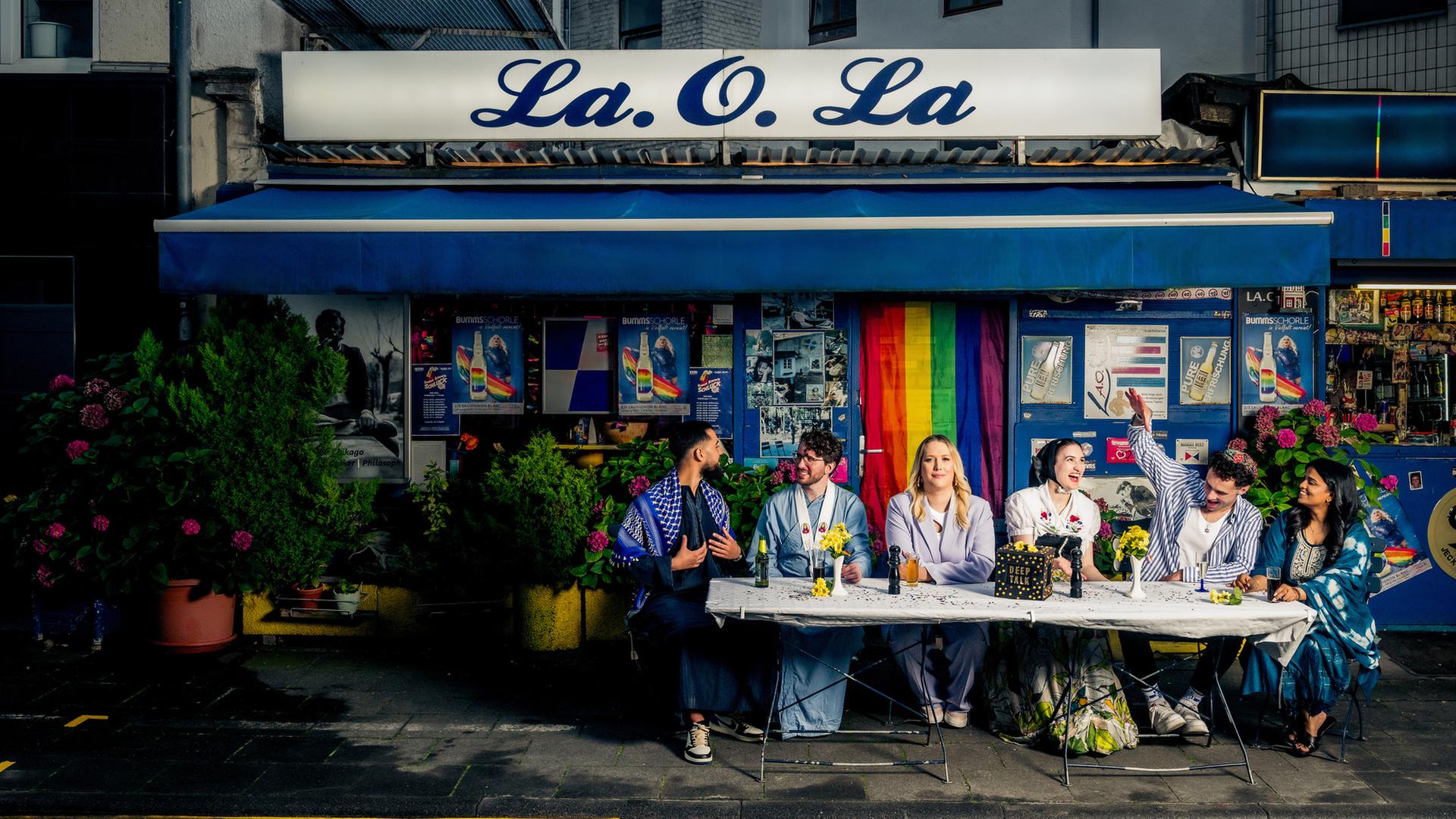 "37°Leben: Göttlich gute Freunde: Zwischen Himmel und Hä?": Sechs Menschen sitzen vor einem Kiosk. Fünf davon sind religiös und sitzen jeweils in ihrer traditionellen Kleidung am Tisch. In der Mitte sitzt eine Atheistin in Alltagskleidung. Vor ihnen auf dem Tisch steht eine Box mit der Aufschrift "Deep Talk".
