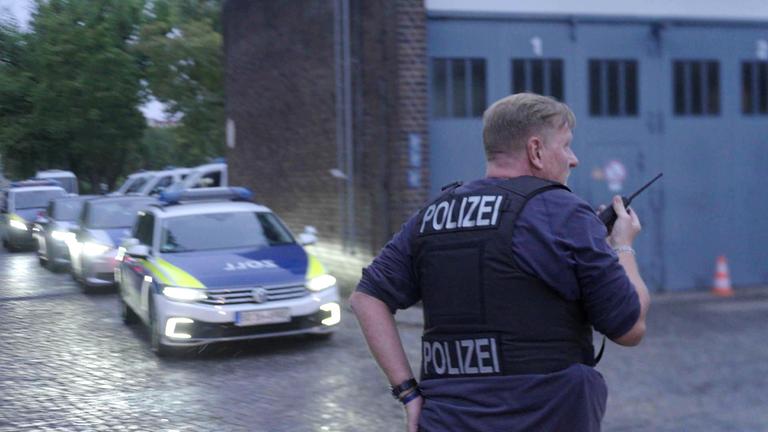 "Kriminelle Clans in Deutschland - Ist der Staat machtlos?": Polizist steht mit dem Rücken zur Kamera auf einer Straße und spricht in ein Funkgerät. Hinter ihm fährt eine Kolonne von Polizeifahrzeugen die Straße entlang.