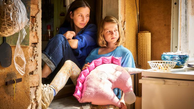 "Familie is nich": Tilda (Luise Landau) und Anne (Meret Becker) sitzen in einem alten Fensterrahmen. Putz bröckelt von der gelb gestrichenen Wand. Tilda hat ein großes Kuschelkissen in Form eines Einhornkopfs auf dem Schoß und schaut zur Seite. Anne schaut liebevoll zu Tilda.
