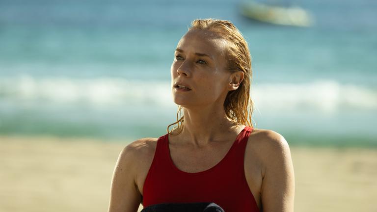 "Visions - Tödliches Verlangen": Halbnah: Estelle (Diane Kruger) steht in einem roten Badeanzug am Strand. Vor der Brust hält sie ein dunkles Handtuch, sie blickt fragend landeinwärts, im Hintergrund liegt das Meer.