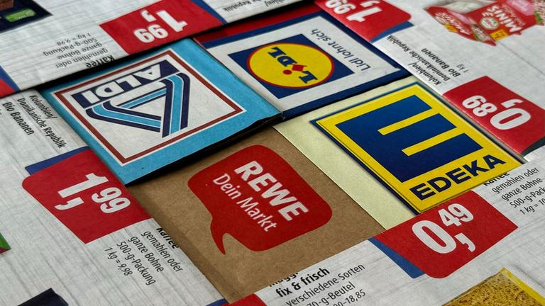 "Rewe, Edeka und Co. - Wer verdient an unseren Lebensmitteln?": Logos und Prospekte von Edeka, Rewe, Aldi und Lidl liegen eng beisammen.