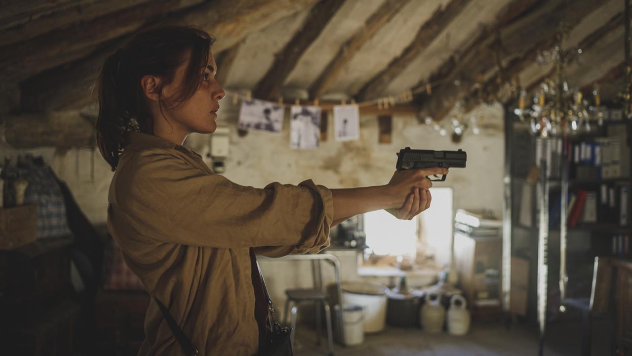 "Blutroter Sommer": Mit der Pistole in Anschlag steht Kommissarin María Ruiz (Marta Nieto) in einem düster wirkenden Lagerraum mit verschiedenen, unscharf abgebildeten Gegenständen im Hintergrund.